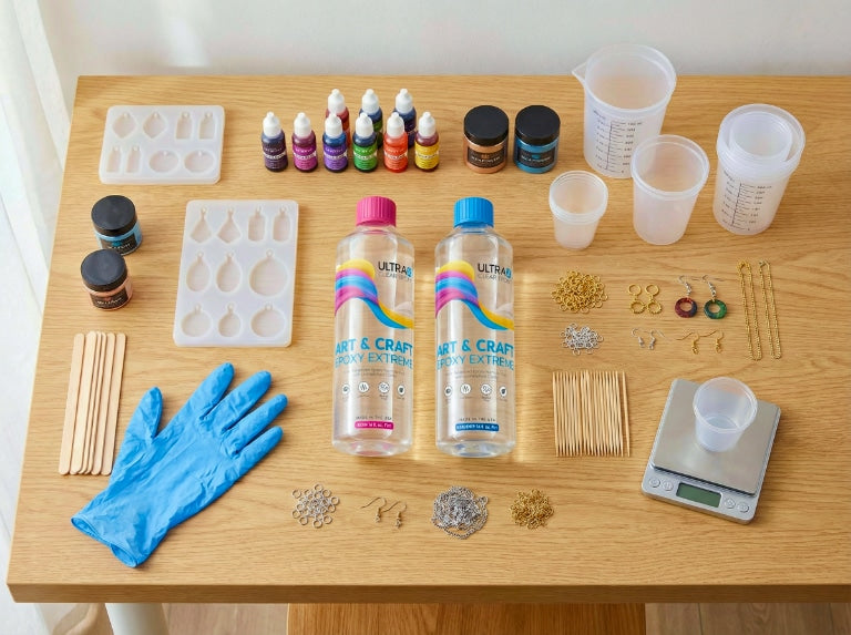 Flat lay of resin jewelry supplies on a wooden table, including silicone molds, UltraClear Art & Craft Epoxy bottles, pigments, mixing cups, gloves, stir sticks, findings, and a digital scale.