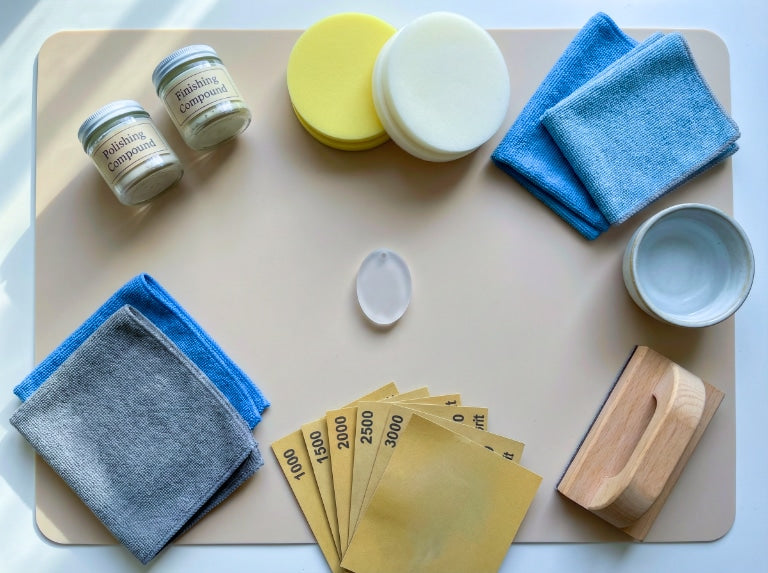 Flat-lay of resin-polishing supplies on a silicone mat, including compounds, foam pads, microfiber cloths, fine-grit sandpapers, bowl, and sanding block.