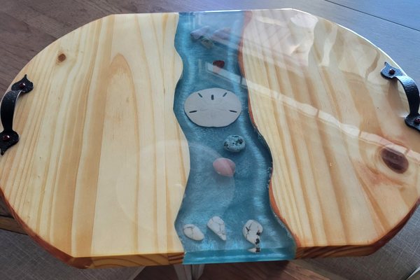 An epoxy river tray with an ocean theme. The river vein was made using UltraClear Art & Craft Epoxy and features a blue-tint and a variety of seashell embedments. The rest of the tray is made of wood and has two metal handles for carrying.