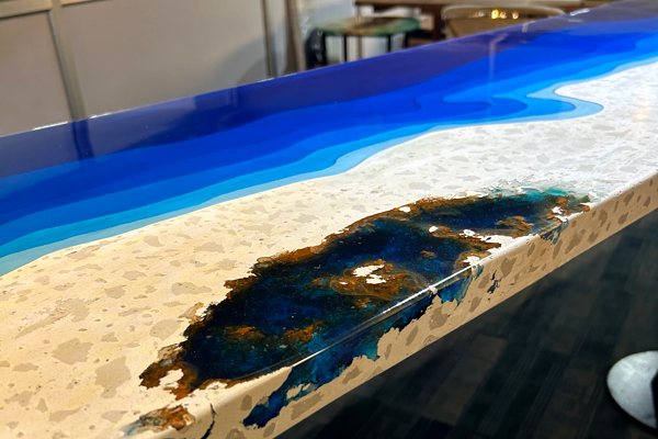 An artistic epoxy table top featuring a quartz countertop substrate with a filled-in space of dark-blue pigmented epoxy and a long, layered section of blue translucent epoxy.