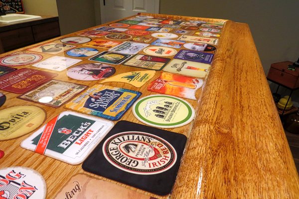 An epoxy bar top with a variety of beer-brand coasters embedded within the UltraClear Bar & Table Top Epoxy finish. The finish is crystal clear, allowing the coasters and underlying wood to be crisply visible.