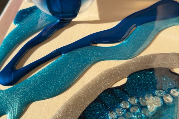 An in-progress photo of an artistic epoxy project. Light blue streak of pigmented epoxy are curing on a wooden tray mold, and dark blue epoxy is being poured in between them.