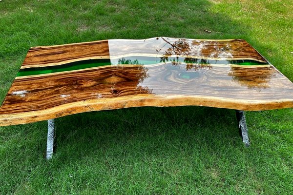 An epoxy river coffee table with a green translucent river vein between two live-edge wooden slabs. It's standing on the grass for display and the reflection of the sky can be seen in its glossy UltraClear Bar & Table Top Epoxy topcoat.