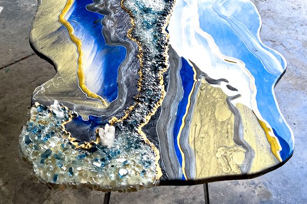A geode-style decorative epoxy coffee table. It features blue and golden-white epoxy pigmented layers, crushed glass embedments, and strip of a metallic material in an unusual shape and abstract design.