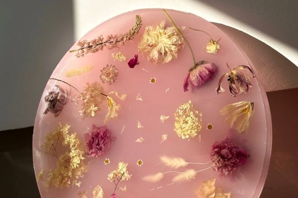 A work of molded resin art consisting of a round opaque pink epoxy layer with various dried flowers and plants embedded within.