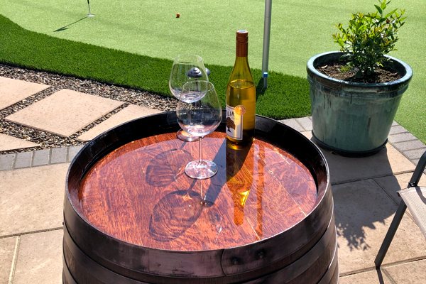 An outdoor epoxy surface consisting of a barrel with a wooden top that has been coated with UltraClear Bar & Table Top Epoxy. Atop the barrel are two wine glasses and a sealed bottle of wine.