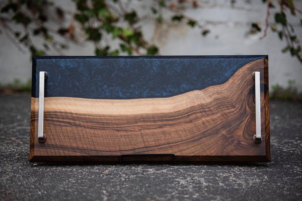 An epoxy serving tray with a deep blue river vein bonded to a piece of wood. It has two metal handles and is positioned for display.