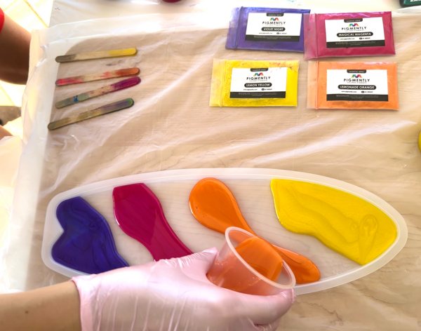 Orange-pigmented epoxy resin being poured into a resin mold between two other small batches of pigmented resin, as part of a resin art project.