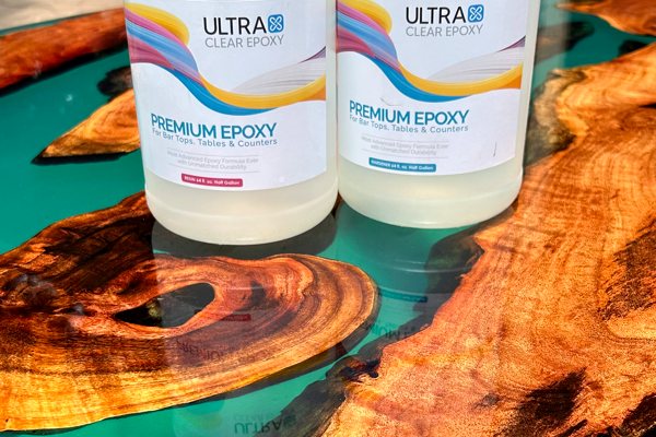 A large round epoxy river table with translucent teal river veins and clear topcoat of UltraClear Bar & Table Top Epoxy. On top of the table are two bottles of UltraClear Bar & Table Top Epoxy.
