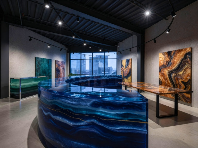 A cinematic showroom with curved blue, green, and amber epoxy surfaces glowing under spotlights, their glassy finishes reflecting light against a modern industrial backdrop with tall windows.