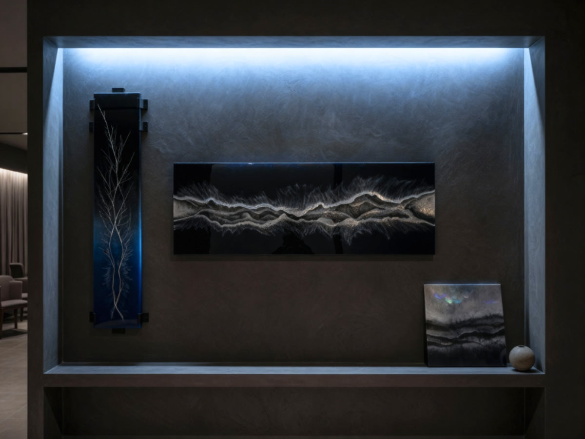 A dark-gray alcove displaying three black epoxy artworks with silver and white highlights, softly lit by blue perimeter lighting, with a small stone accent beside the smallest piece.