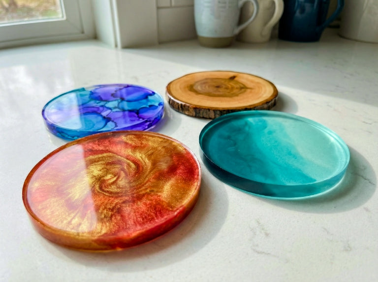 Four finished epoxy resin coasters resting flat on a light kitchen countertop: a teal translucent coaster, a red-and-gold metallic swirl coaster, a blue-purple alcohol-ink style coaster, and a wood slice coaster sealed with clear epoxy.