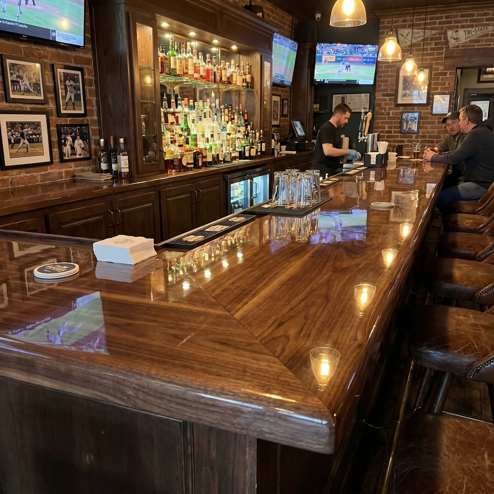 Glossy epoxy-coated wood bar top in a busy brick-walled sports bar with patrons, bartender, and illuminated liquor display.
