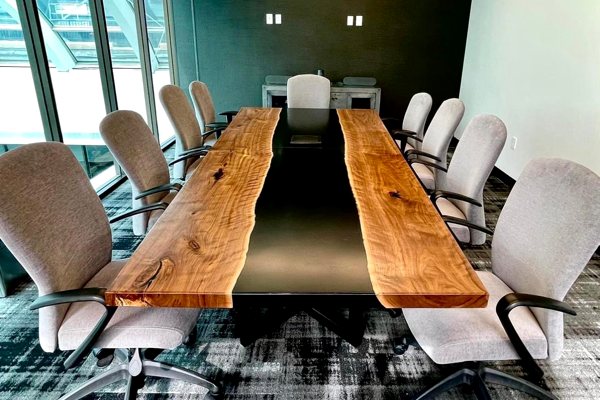 A large epoxy river table with an extra-wide black-tinted vein of UltraClear Deep Pour Epoxy between two long wooden slabs. It is resting in a conference room and surrounded by fabric office chairs.