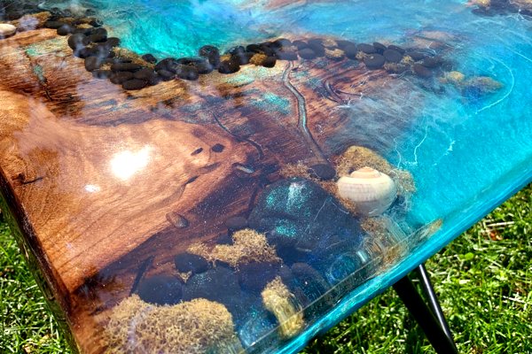 An artistic resin river table made from a very thick, tinted epoxy coating. It features a variety of sea-themed embedments.
