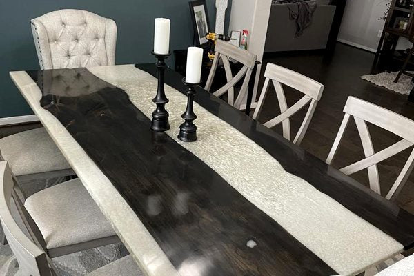 A large epoxy river dining table. It features black-painted wooden slabs with a thick, wide river vein of UltraClear Deep Pour Epoxy in between. The vein has been colored with a white pigment.