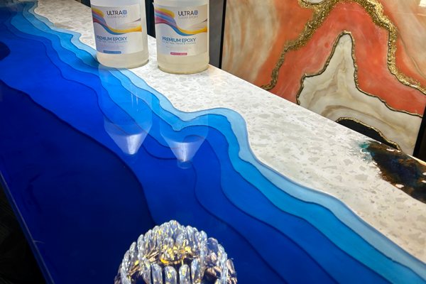 An artistic epoxy table top. Multiple blue-tinted UltraClear Deep Pour Epoxy layer depict a water scene against a quartz substrate, and the whole table has been sealed with UltraClear Bar & Table Top Epoxy.