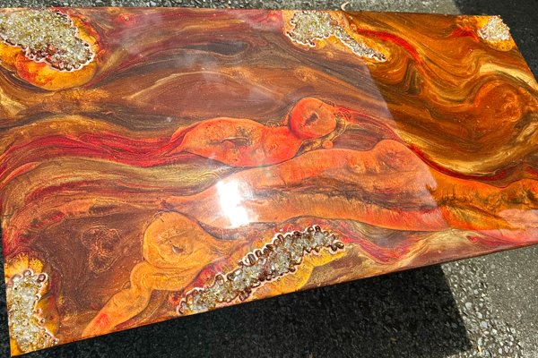 An artistic epoxy coffee table featuring several tinted UltraClear Art & Craft Epoxy streaking layers of orange and brown tones. There are geode-like crystals embedded into the finish at sporadic locations near the edges.