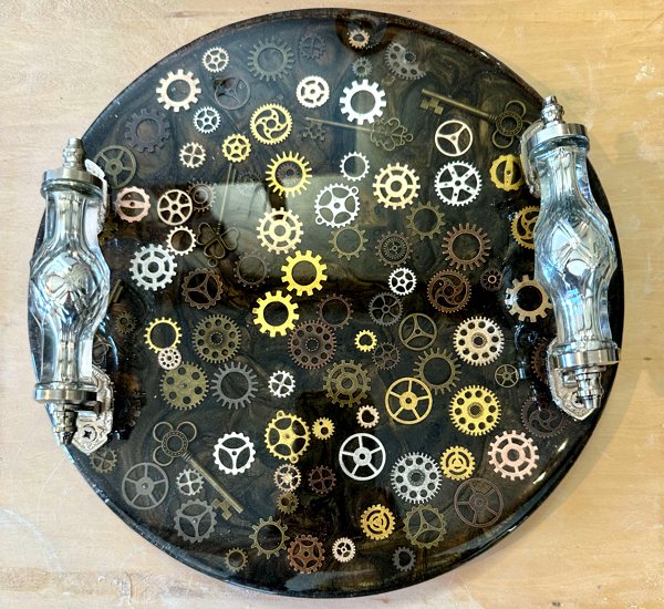 An intricate epoxy resin tray with metallic handles and a clear epoxy base filled with clockwork-themed embedments such as tiny gears and cogs. It was made with UltraClear Art & Craft Epoxy.