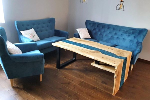 A living room with a large, elaborate epoxy river coffee table. The table features a turquoise-pigmented river vein and is surrounded by turquoise fabric furniture.