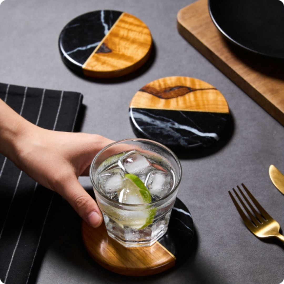 Hand holding a glass of iced drink with lime on a wooden coaster, surrounded by other coasters and cutlery.